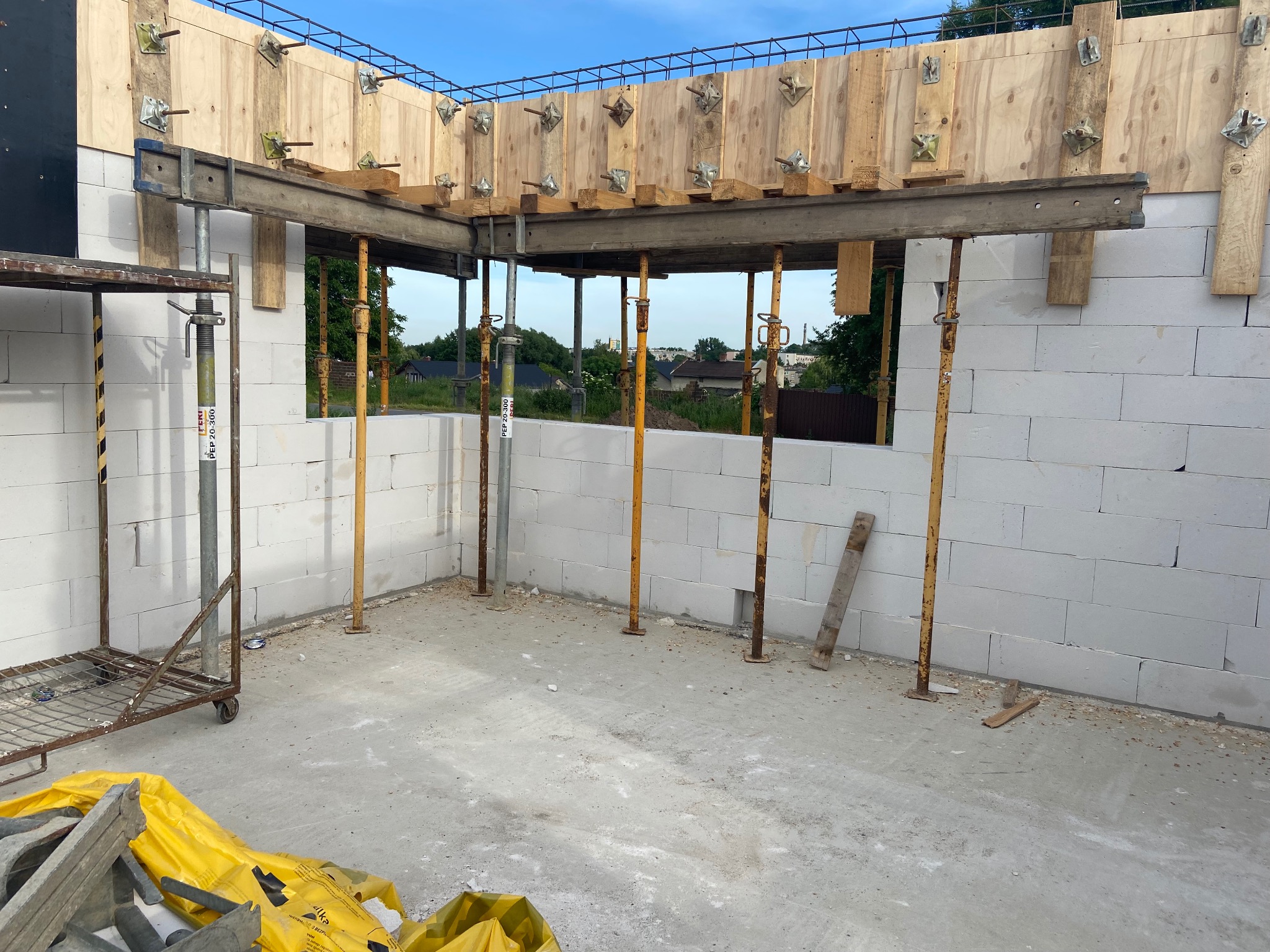 Wnętrze w trakcie budowy domu z białych bloczków, widoczne żółte stemple podtrzymujące deskowanie, metalowy wózek z narzędziami w rogu, w tle zieleń drzew.