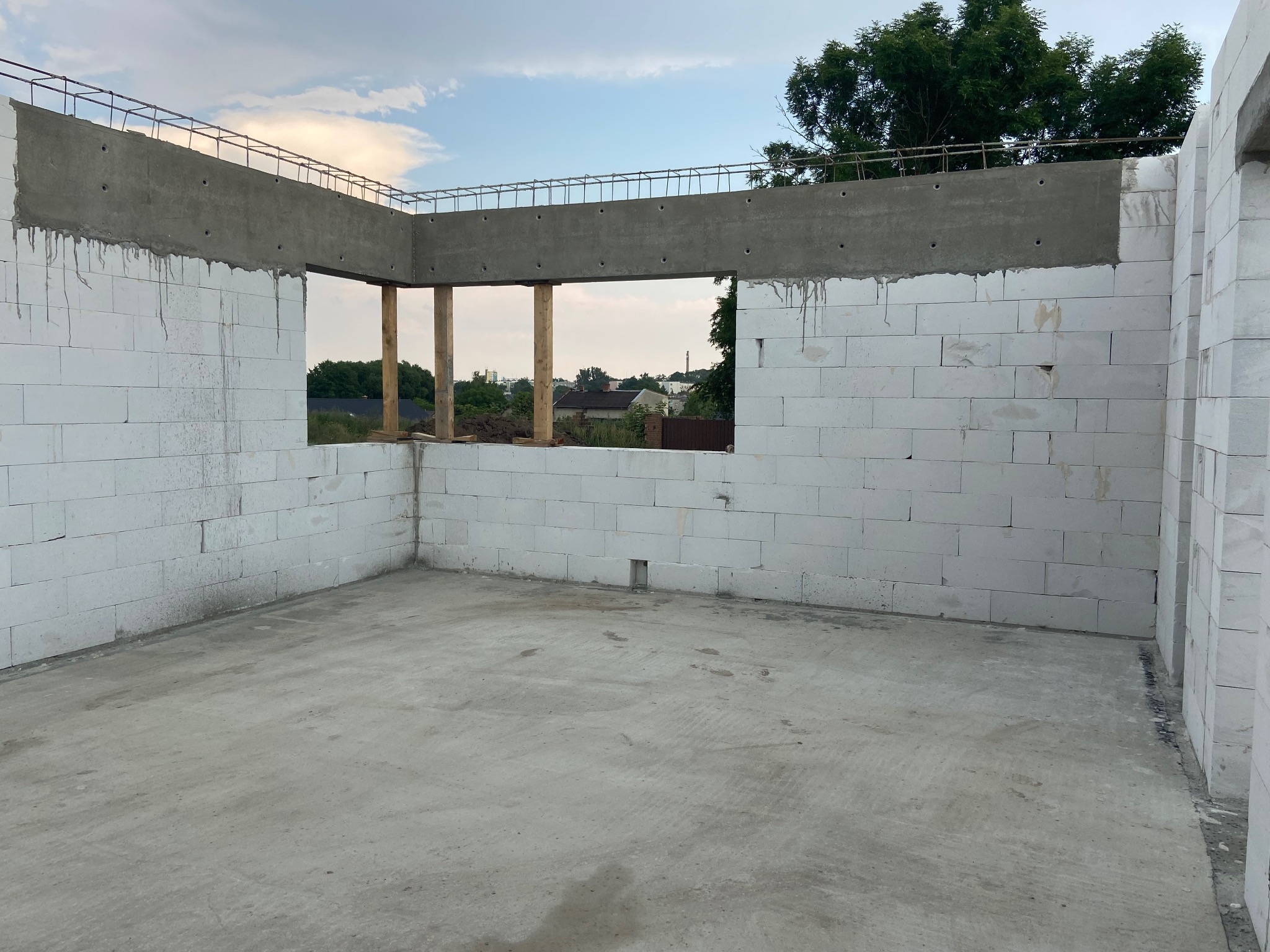 Wnętrze surowego domu w trakcie budowy, ściany z białych bloczków, betonowy strop z wystającą zbrojeniem, widoczne drewniane podpory okna, w tle zieleń drzew i zabudowa.
