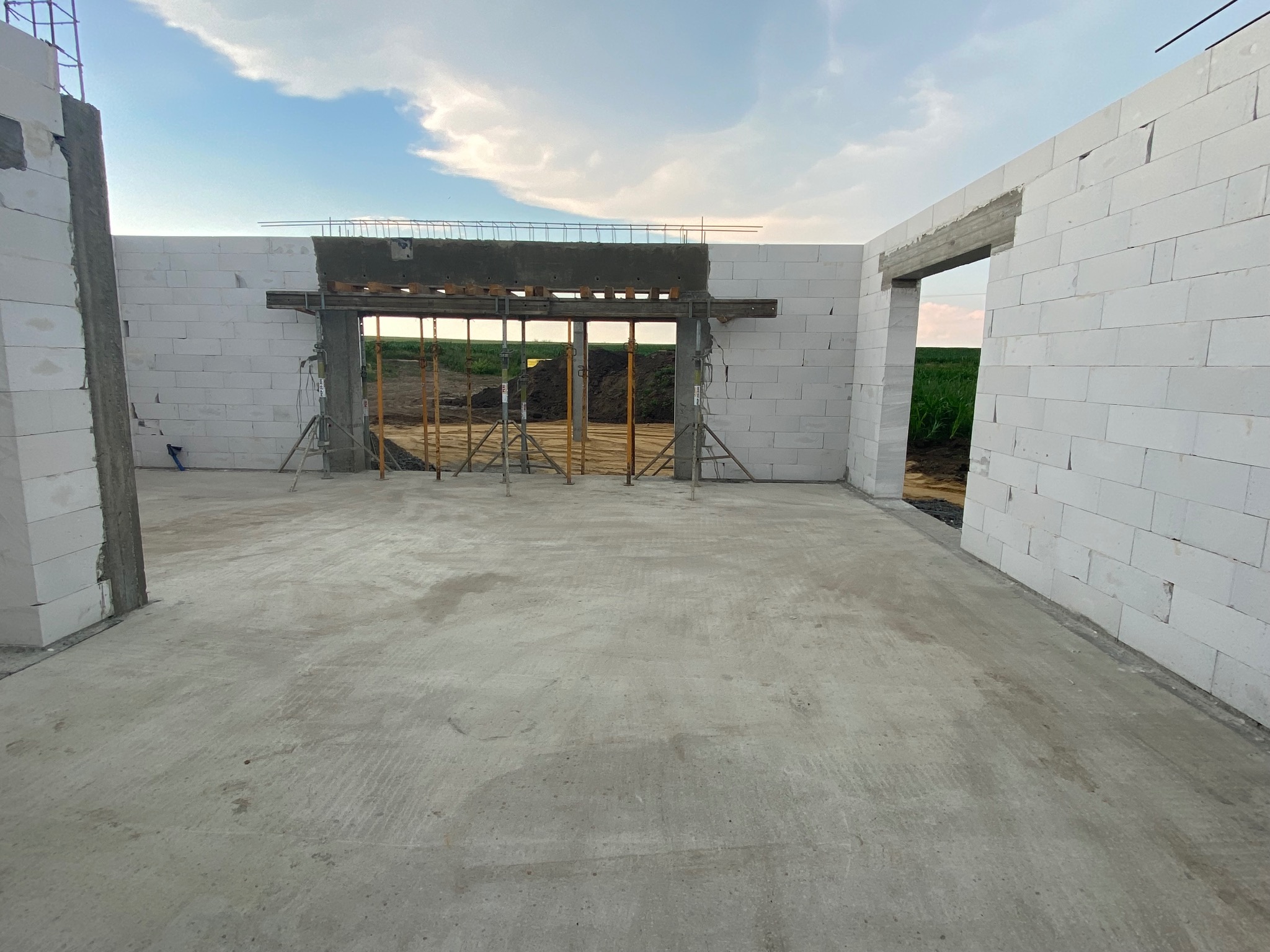 Wnętrze domu w trakcie budowy, ściany z białych bloczków, betonowa wylewka, widoczne szalunki i podpory pod nadprożem, w tle zielone pole.