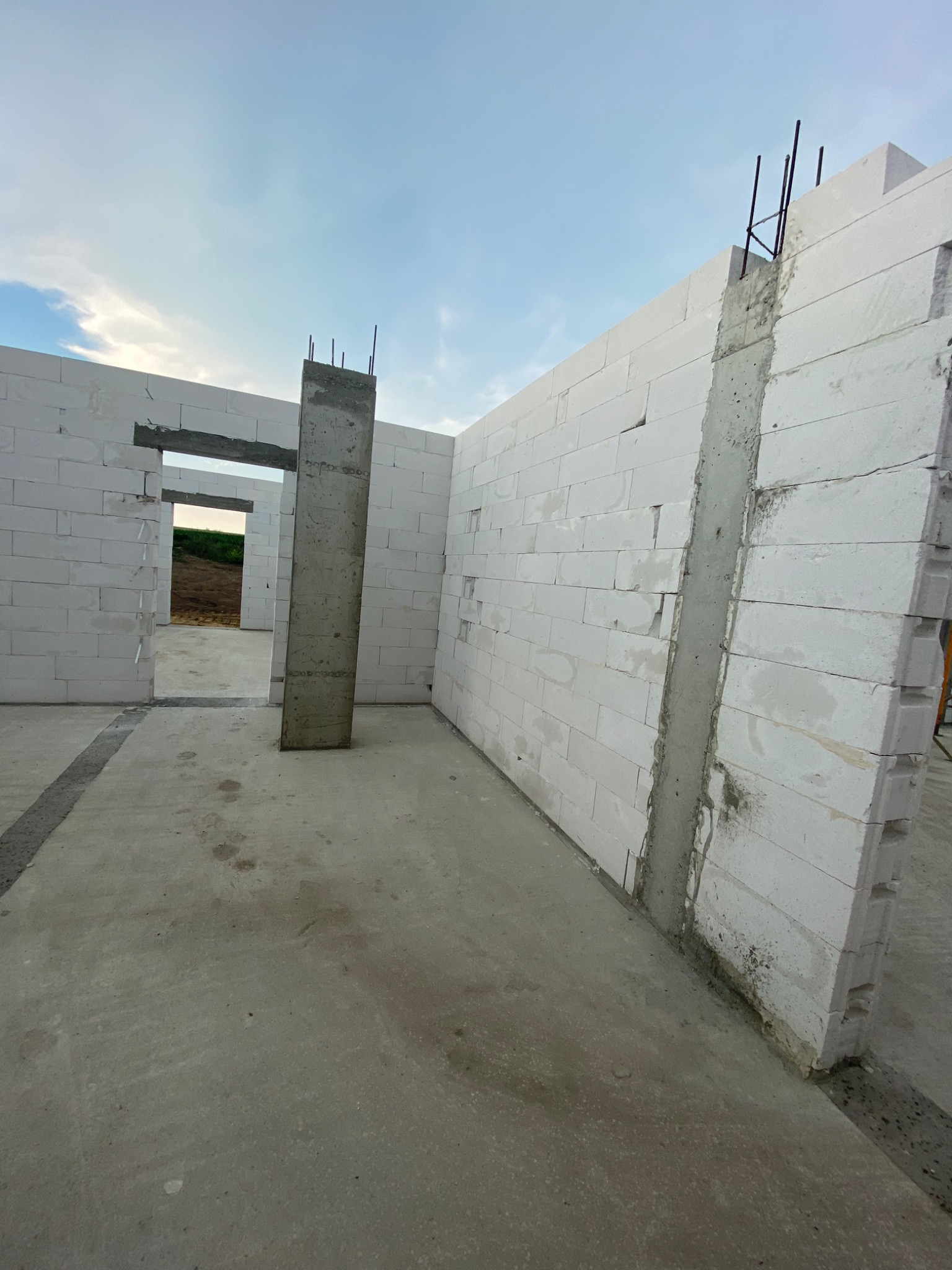 Wnętrze surowego domu w trakcie budowy, widoczne ściany z białych bloczków z betonu komórkowego, zbrojony betonowy filar i wylewka podłogowa. Na zewnątrz widoczny fragment terenu i niebo.