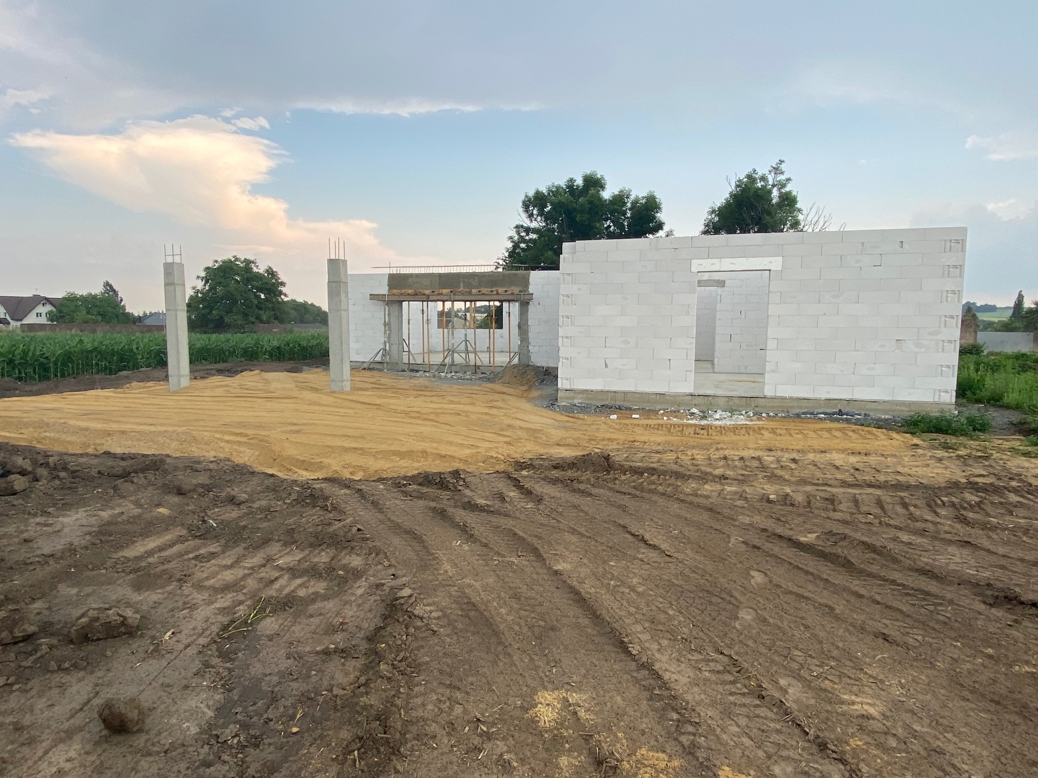 Budowa domu z betonu komórkowego w trakcie wznoszenia ścian, widoczne zbrojone słupy i szalunki nadproży, teren wokół wyrównany piaskiem, w tle pole uprawne.