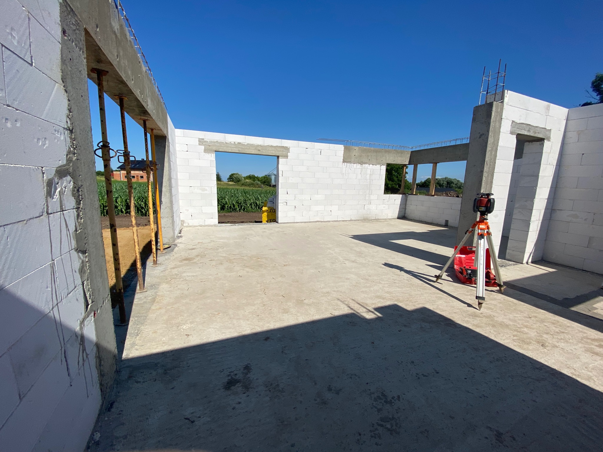 Wnętrze surowego domu murowanego w trakcie budowy, ściany z białych bloczków, widoczne podpory szalunkowe, poziomica laserowa na statywie, w tle pole kukurydzy i budynek mieszkalny, błękitne niebo.