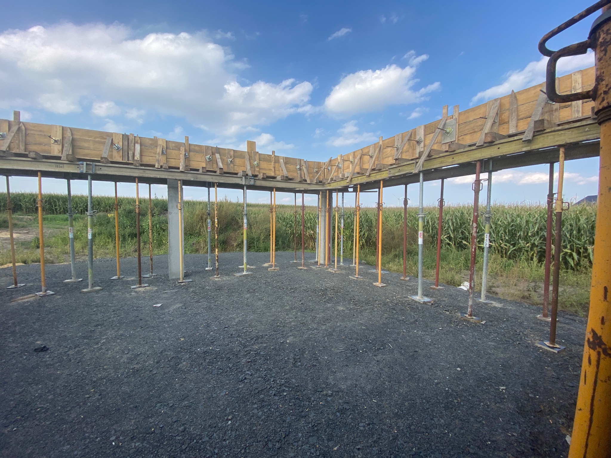 Szare podłoże z licznymi metalowymi podporami i drewnianym szalunkiem w tle pola kukurydzy pod błękitnym niebem z chmurami, widoczny element żółtej podpory po prawej stronie kadru.