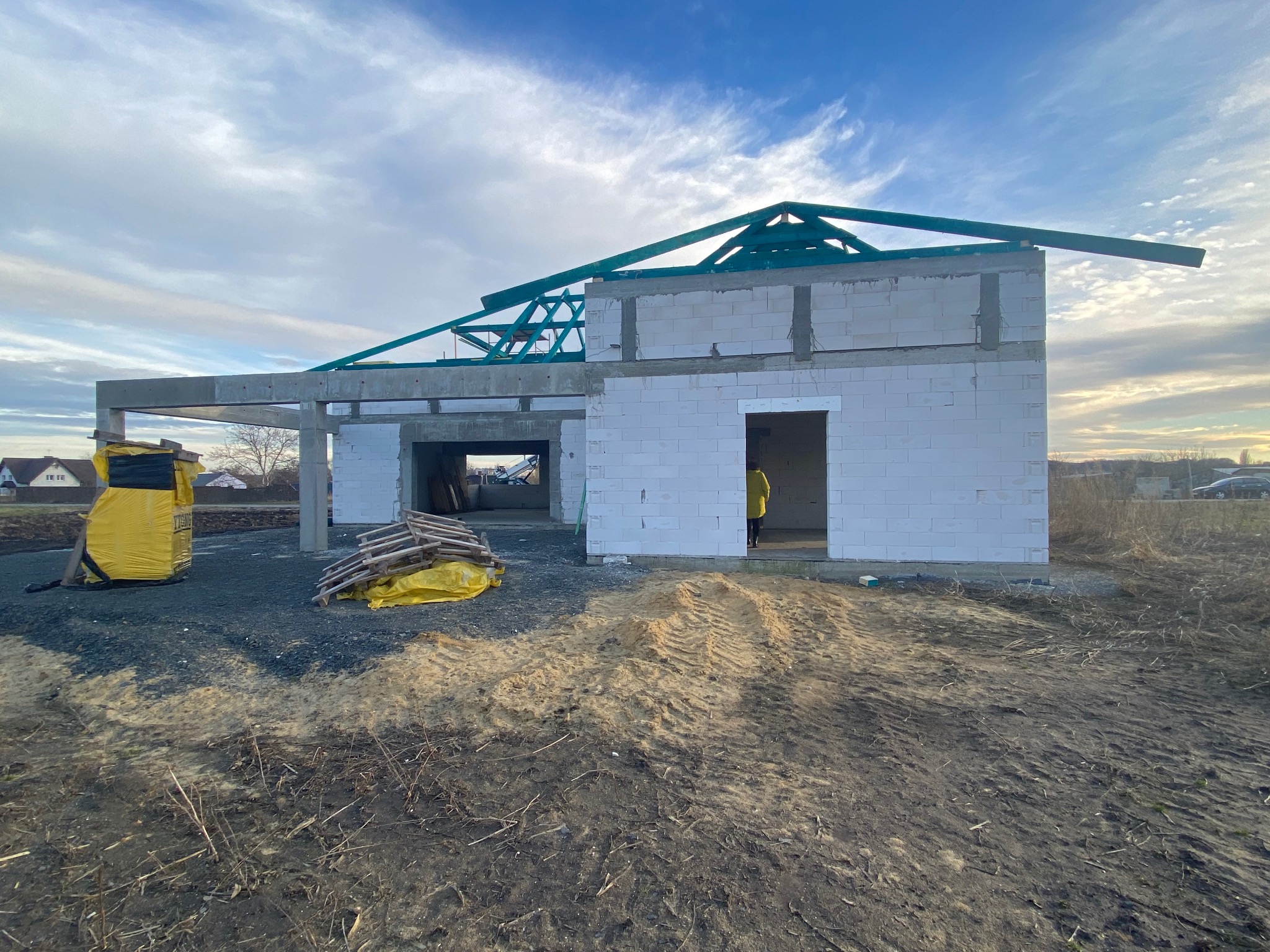 Budowa domu w stanie surowym otwartym z betonu komórkowego, widoczne elementy więźby dachowej, na pierwszym planie ziemia z wyraźnymi śladami opon, w tle zabudowa jednorodzinna.