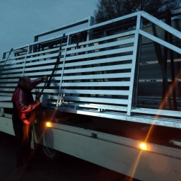 Eko Płot Systemy Ogrodzeniowe Krzysztof Łasiński - Mężczyzna zabezpiecza metalowe elementy konstrukcyjne ogrodzenia na platformie transportowej przy słabym oświetleniu.