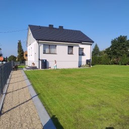 Eko Płot Systemy Ogrodzeniowe Krzysztof Łasiński - Nowoczesny dom jednorodzinny z szarym ogrodzeniem panelowym, żwirową ścieżką i rozległym trawnikiem w słoneczny dzień.