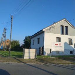 Eko Płot Systemy Ogrodzeniowe Krzysztof Łasiński - Nowoczesny dom z szarą elewacją i dachem, otoczony siatką ogrodzeniową, z widoczną tablicą informacyjną o dofinansowaniu remontu dróg gminnych ze środków rządowych.