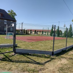 Eko Płot Systemy Ogrodzeniowe Krzysztof Łasiński - Otwarta furtka z szarej siatki ogrodzeniowej na betonowej podmurówce, z widokiem na boisko sportowe i budynek w tle, w słoneczny dzień.