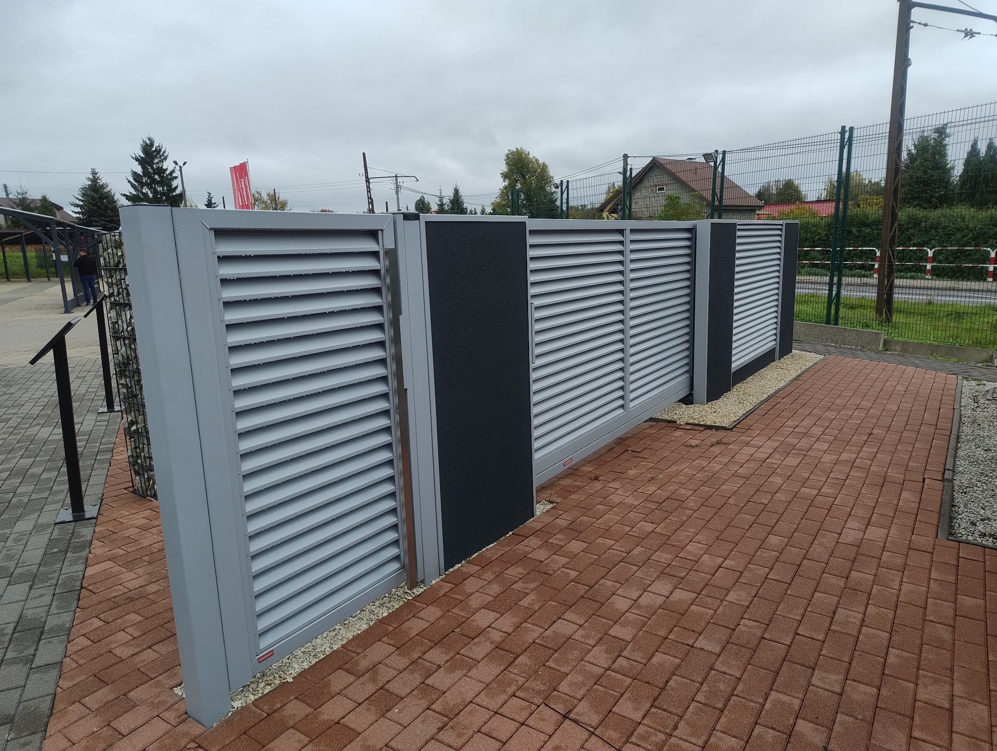 Nowoczesne, szare przęsła z poziomymi lamelami i czarnymi panelami, oddzielone drobnym żwirem od czerwonej kostki brukowej.