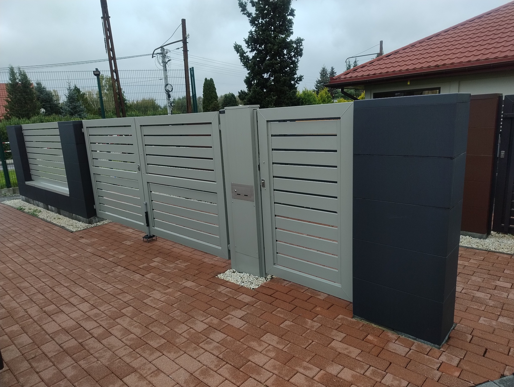 Nowoczesna, pozioma brama panelowa w odcieniach szarości z wbudowaną skrzynką na listy, osadzona na ceglanej kostce brukowej.