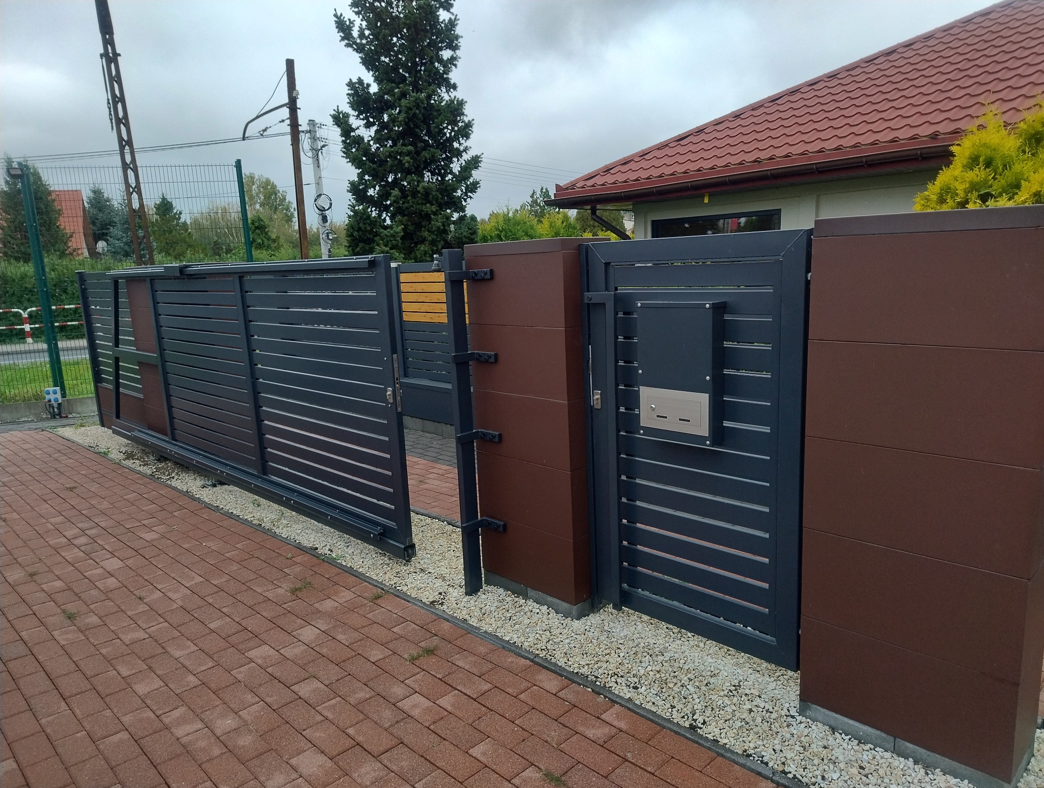 Nowoczesna, grafitowo-brązowa brama wjazdowa z poziomymi panelami, wyposażona w skrzynkę na listy, zamontowana na posesji z kostką brukową i żwirowym obrzeżem.