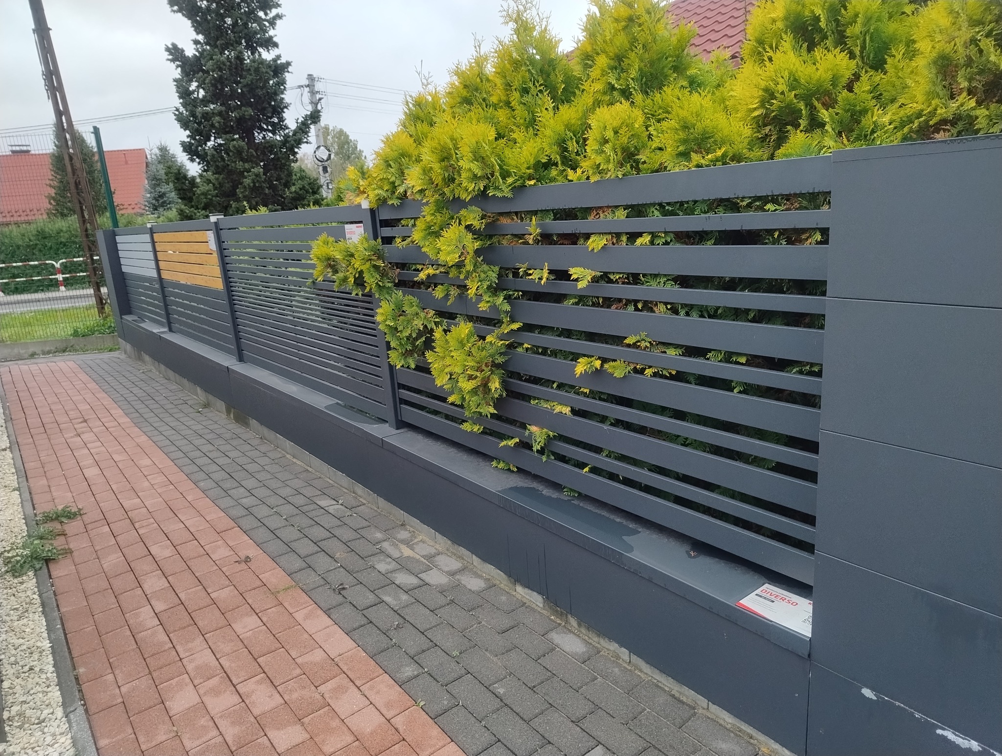 Nowoczesne, poziome, szare ogrodzenie z elementami drewnianymi, częściowo porośnięte zielonymi krzewami, przylegające do dwukolorowej kostki brukowej.