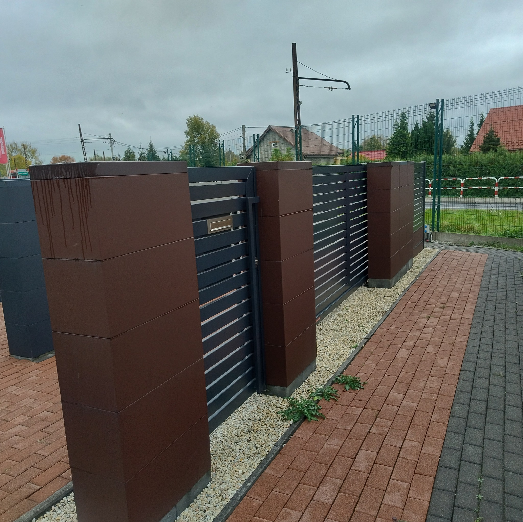 Nowoczesna brama wjazdowa z poziomymi panelami w kolorze grafitowym i brązowymi słupkami, widoczna skrzynka na listy, otoczenie z kostki brukowej i żwiru.