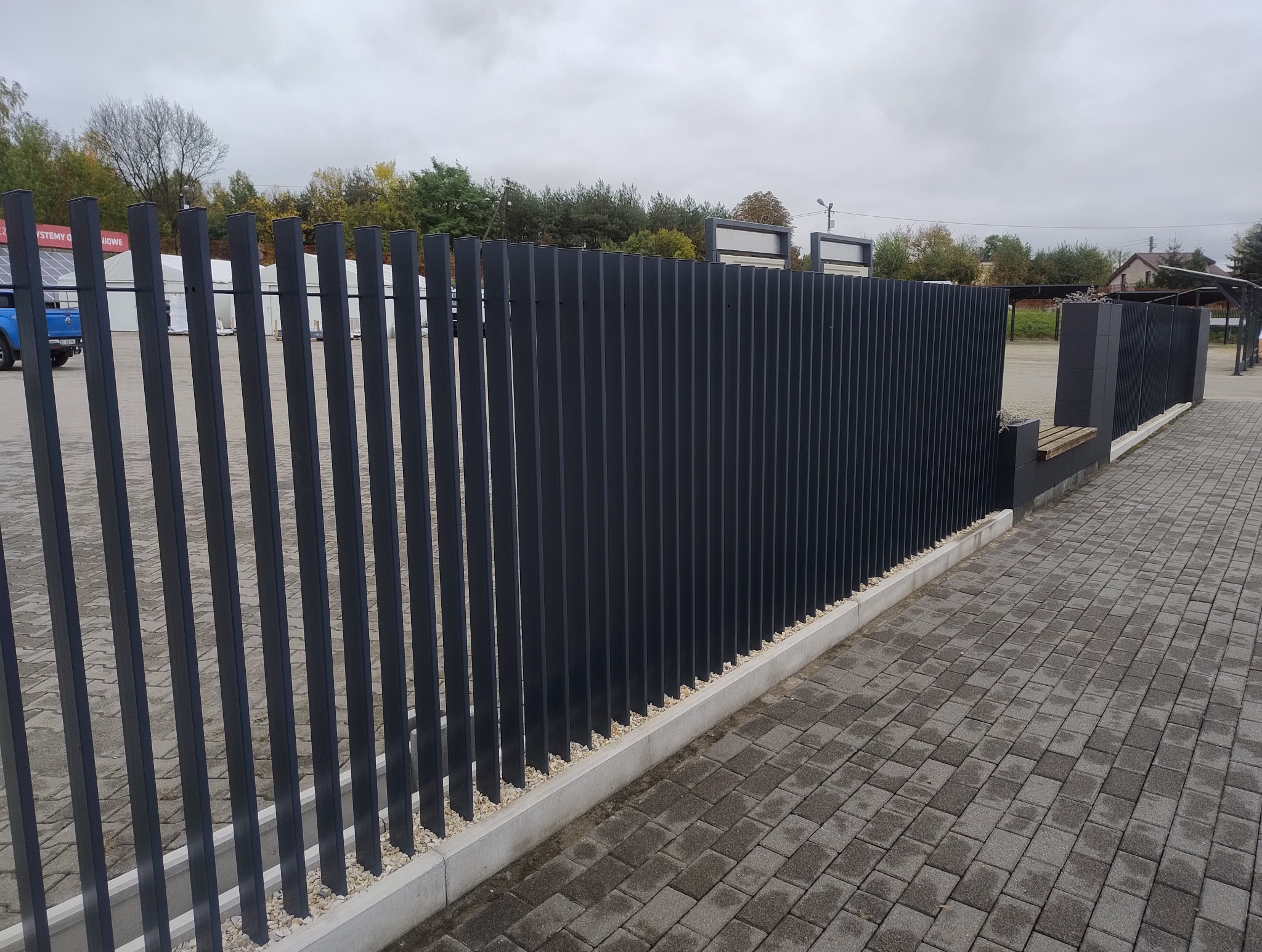 Nowoczesne, pionowe, metalowe przęsła ogrodzeniowe w kolorze grafitowym, zamontowane na betonowej podmurówce z białym grysem, widok z boku na kostkę brukową.