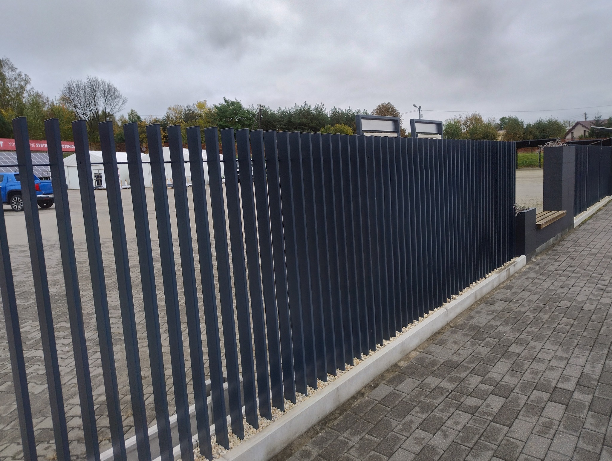 Nowoczesne, pionowe, grafitowe przęsła ogrodzeniowe z metalu, osadzone na betonowej podmurówce z drobnym kamieniem, obok kostka brukowa i element z siedziskiem.