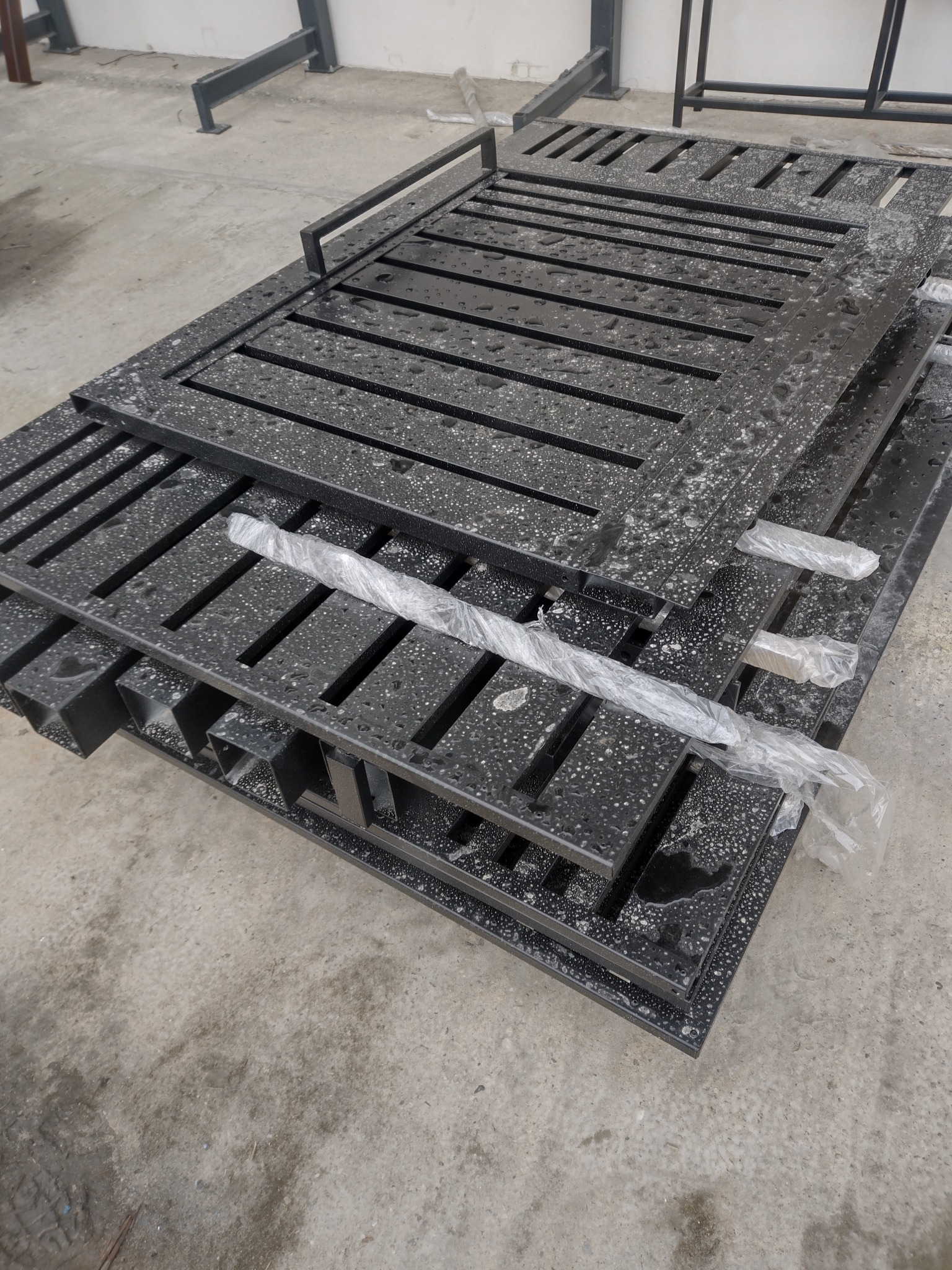 Stos czarnych, metalowych elementów ogrodzeniowych z poziomymi szczelinami, zabezpieczonych folią, leżących na betonowej posadzce w hali.