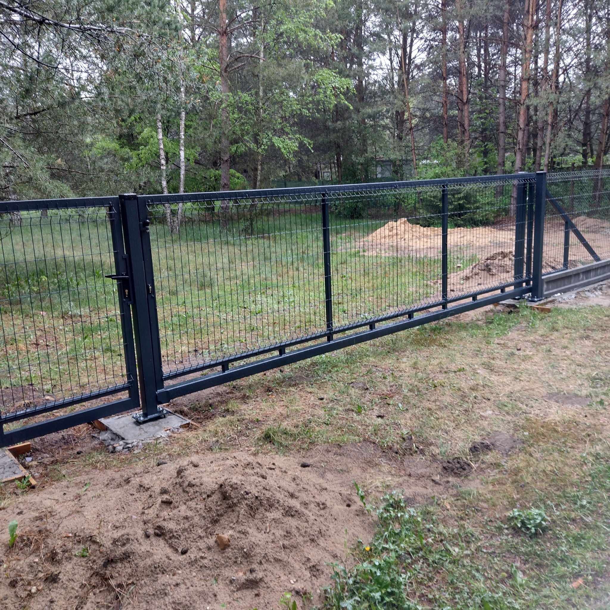 Przesuwna brama z paneli ogrodzeniowych, z widocznym zamkiem i słupkami, zamontowana na betonowej podstawie, na tle leśnego krajobrazu.