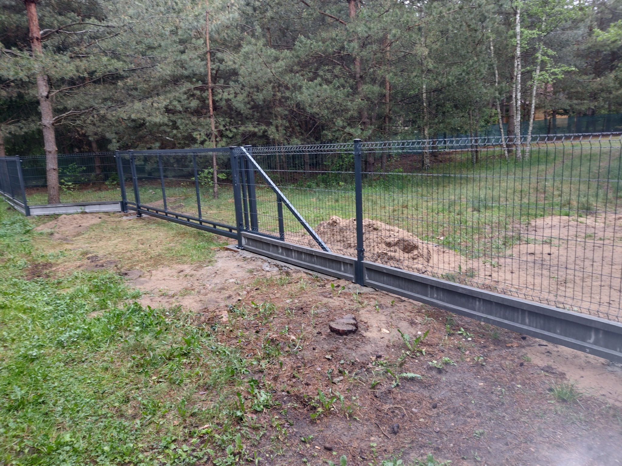 Ciemnoszara, przesuwna brama z siatki panelowej z ukośnym wspornikiem, zamontowana na betonowej podstawie w leśnym otoczeniu.
