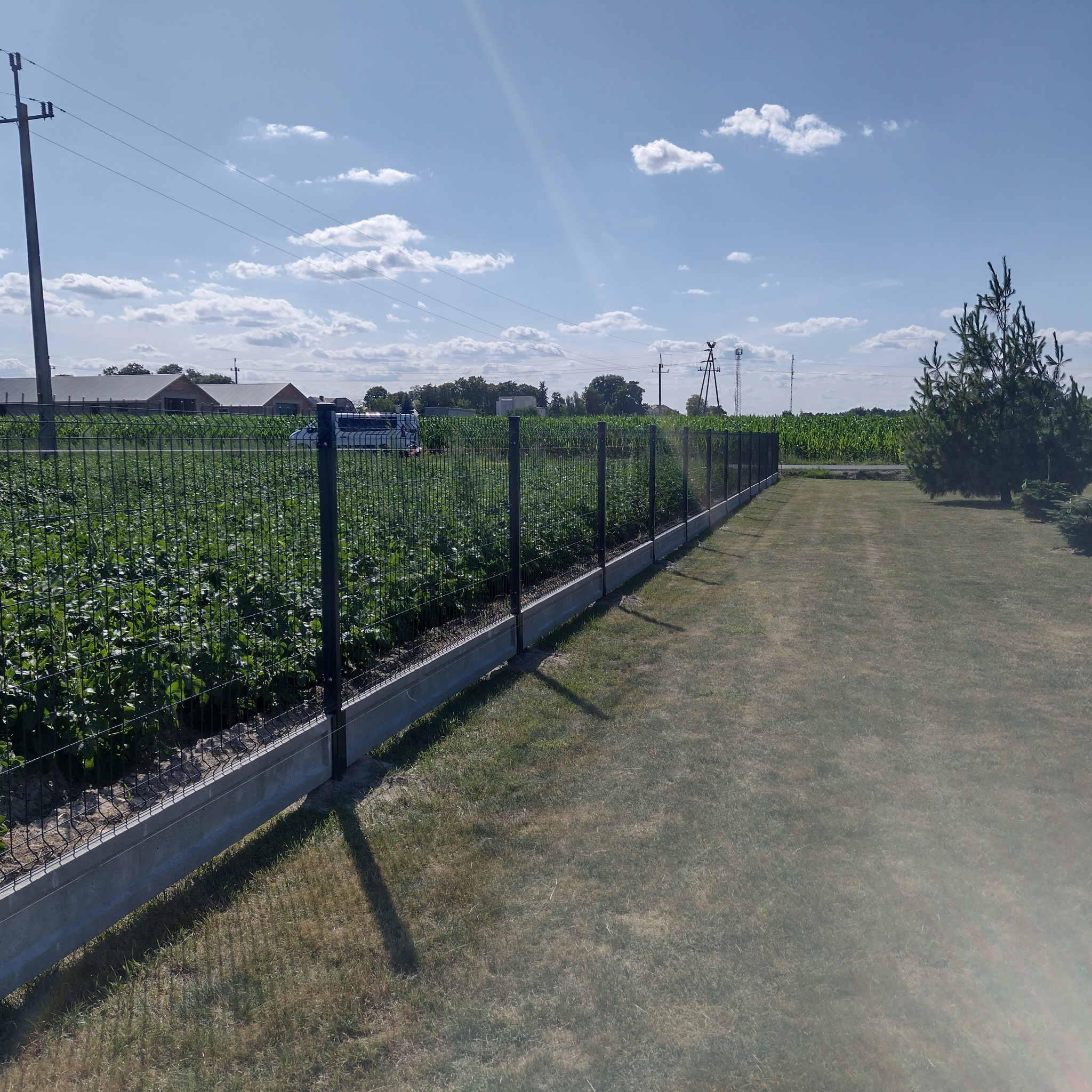 Długi fragment posesji z nowym, czarnym ogrodzeniem z siatki panelowej na betonowej podmurówce, za którym widać pole uprawne i budynki gospodarcze w oddali.