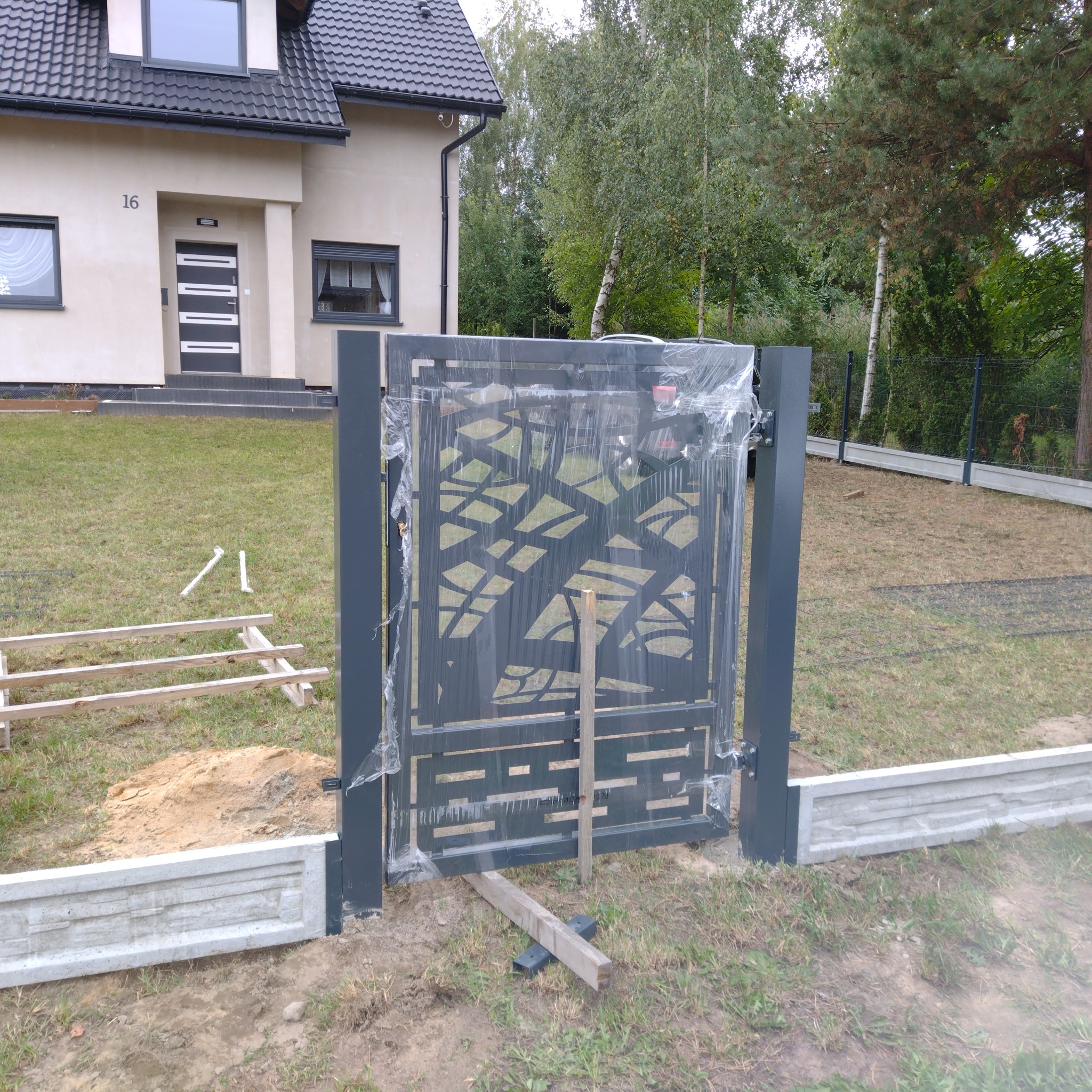 Nowoczesna, metalowa brama z ażurowym wzorem, jeszcze w folii ochronnej, w trakcie montażu przed domem z numerem 16, widoczne elementy konstrukcyjne i podkłady.