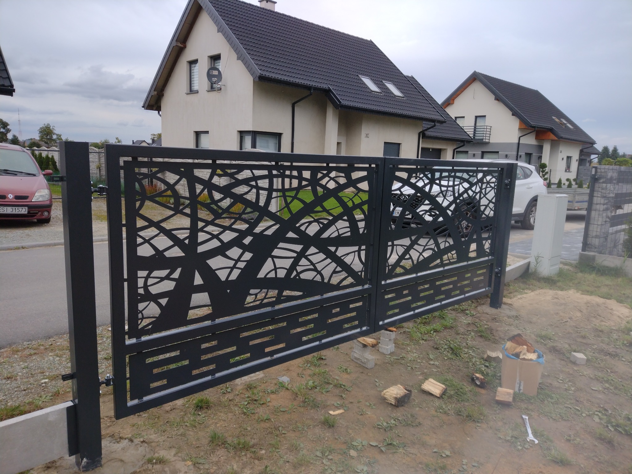 Nowoczesna, czarna brama wjazdowa z ażurowym wzorem na tle dwóch domów jednorodzinnych, widoczne bloczki betonowe i narzędzia sugerujące etap montażu.