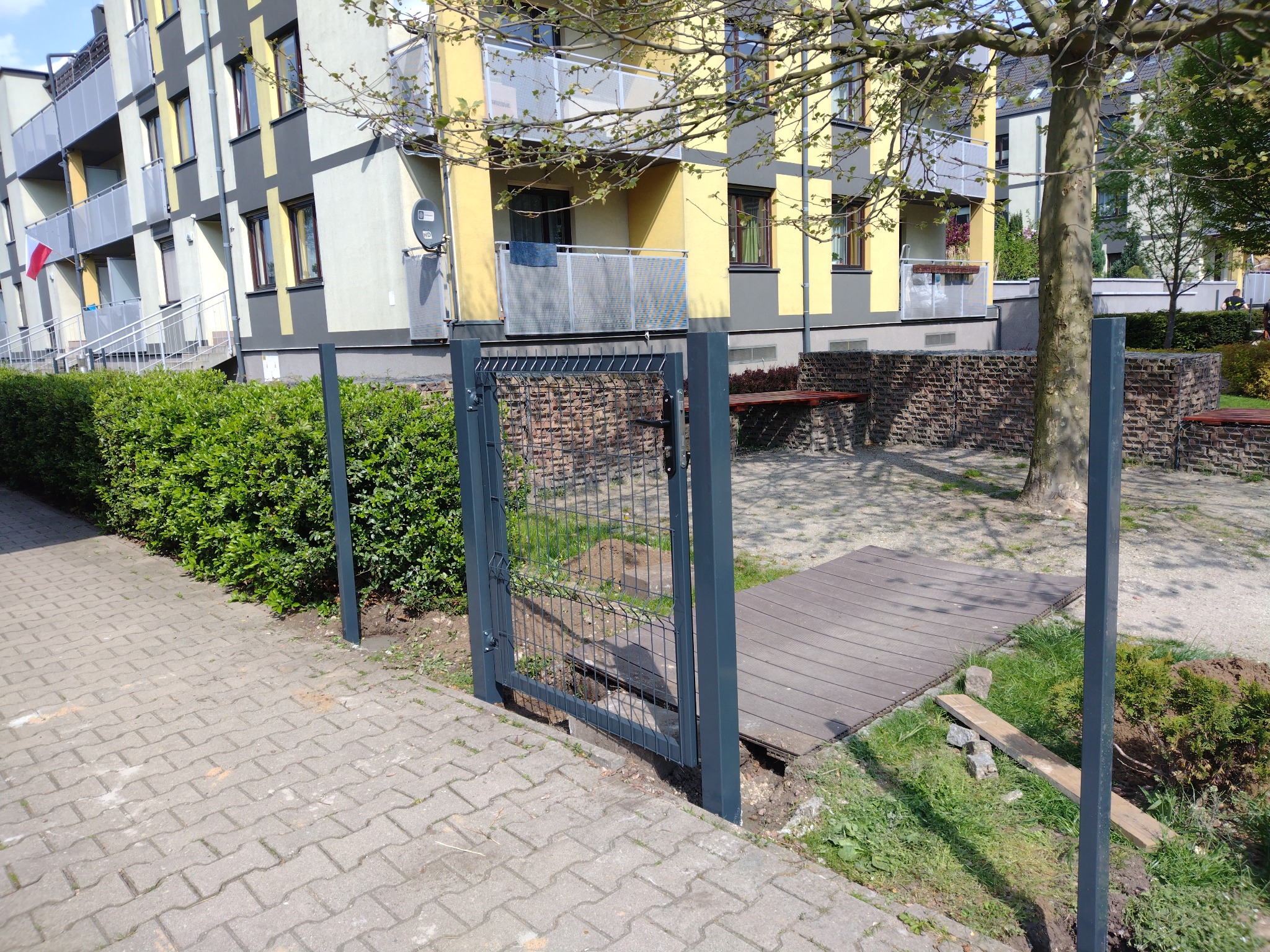 Szara, metalowa furtka z siatki, częściowo zamontowana, widoczne słupki i wykopane dołki w ziemi, na tle bloku mieszkalnego i chodnika z kostki brukowej.