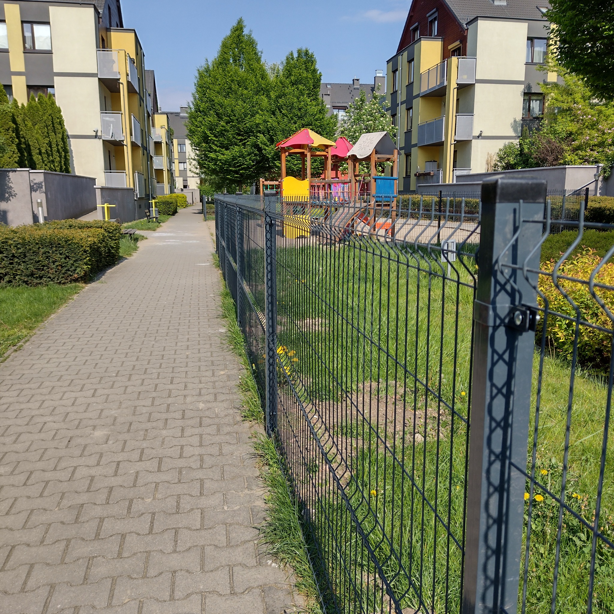 Nowoczesne, grafitowe ogrodzenie panelowe oddzielające chodnik z kostki brukowej od trawnika z placem zabaw w tle, w otoczeniu wielorodzinnych budynków.