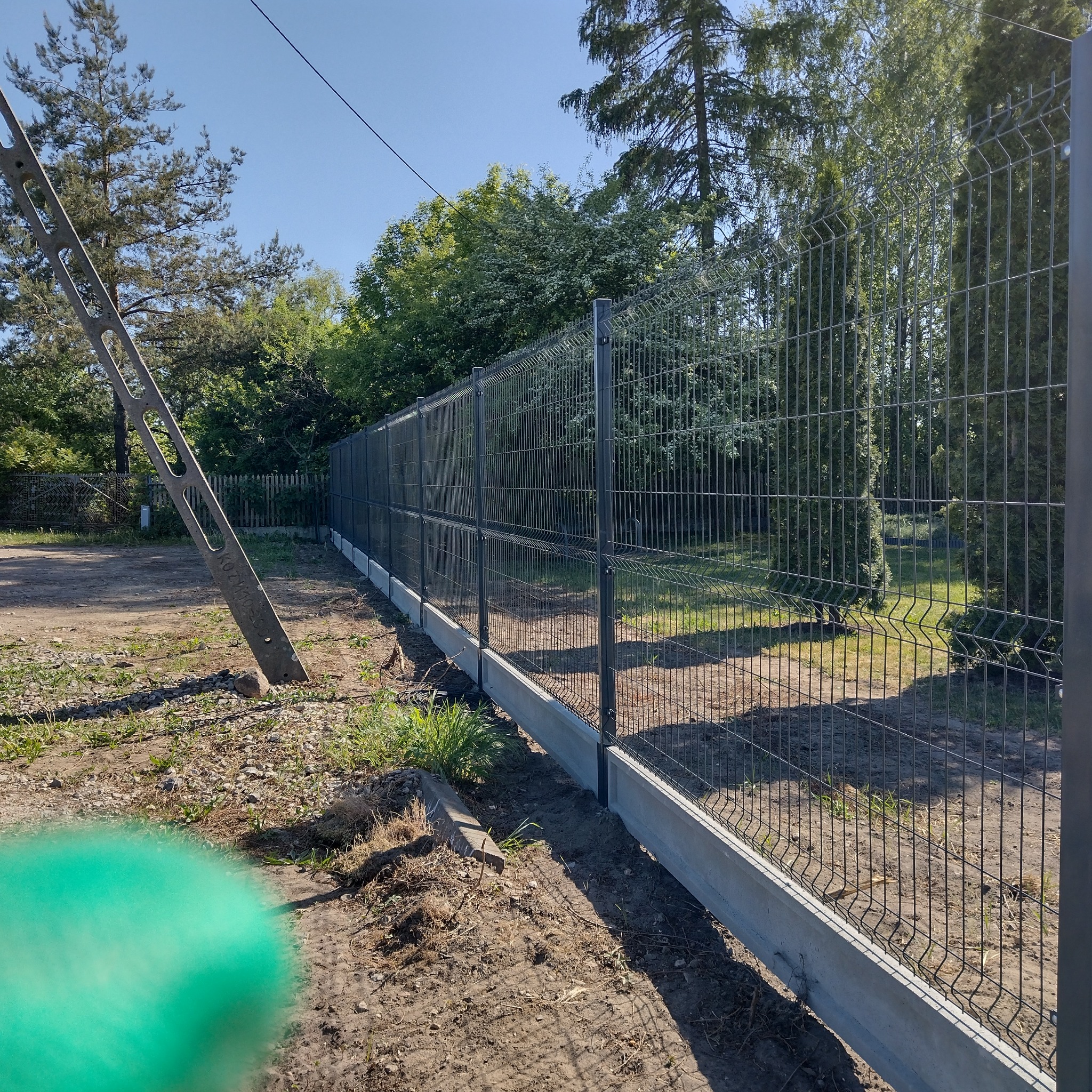 Długi, prosty odcinek ciemnoszarego ogrodzenia z paneli na betonowej podmurówce, widok z boku, z fragmentem słupa energetycznego po lewej stronie i zaroślami w tle.