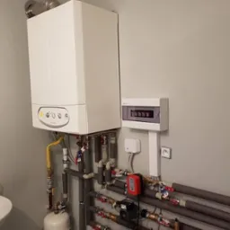 Biały piec gazowy wiszący na szarej ścianie, z podłączonymi rurami miedzianymi i elementami instalacji grzewczej, widoczna skrzynka bezpiecznikowa i mała umywalka z lewej strony.