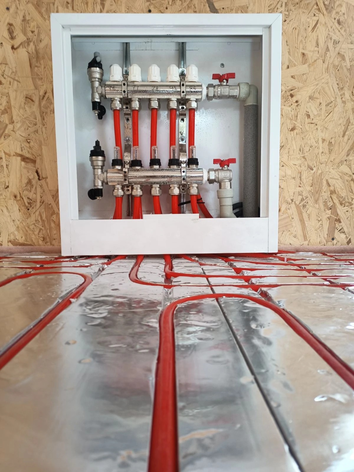 Rozdzielacz instalacji ogrzewania podłogowego w białej szafce, z widocznymi czerwonymi rurami na srebrnej folii izolacyjnej.