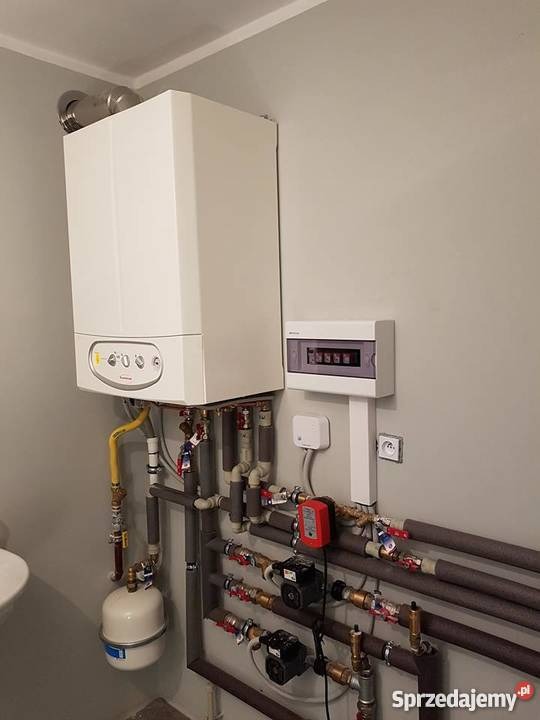 Biały piec gazowy wiszący na szarej ścianie, z podłączonymi rurami miedzianymi i elementami instalacji grzewczej, widoczna skrzynka bezpiecznikowa i mała umywalka z lewej strony.