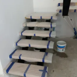 Wewnętrzne schody drewniane na czarnej metalowej konstrukcji, zabezpieczone folią i taśmą malarską podczas prac wykończeniowych, widoczny fragment ściany z wystającymi przewodami elektrycznymi...