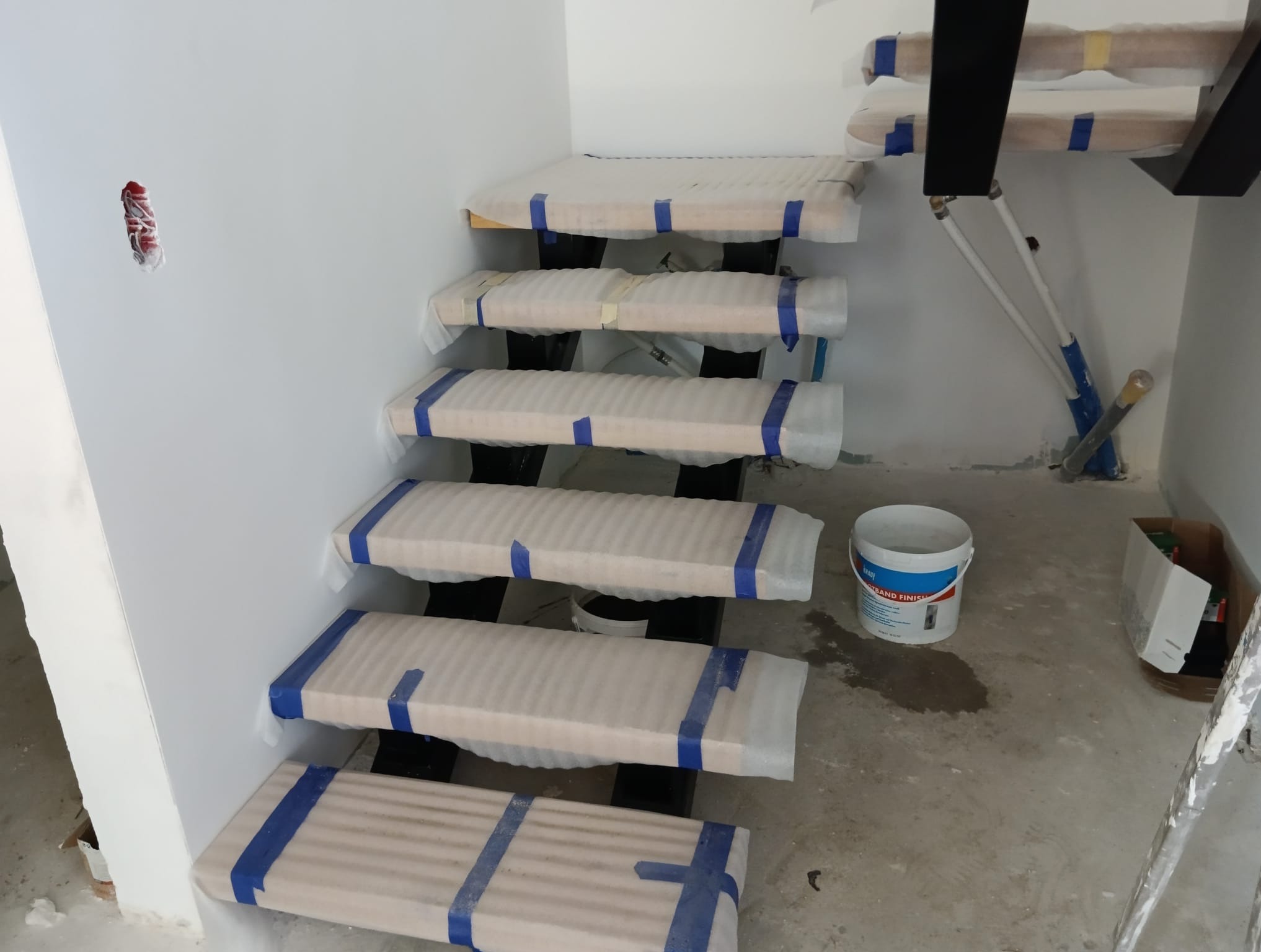 Wewnętrzne schody drewniane na czarnej metalowej konstrukcji, zabezpieczone folią i taśmą malarską podczas prac wykończeniowych, widoczny fragment ściany z wystającymi przewodami elektrycznymi...