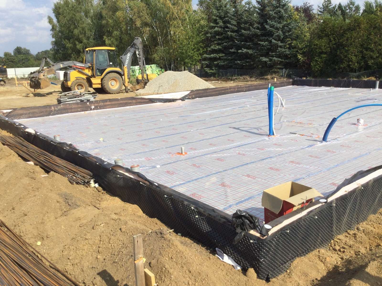 Przygotowany teren pod wylewkę fundamentów z widoczną izolacją pionową, folią z siatką, wystającymi rurami instalacyjnymi oraz żółtą koparką Volvo w tle.