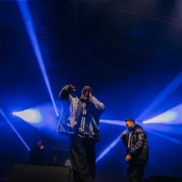 Dwóch wokalistów na scenie oświetlonej niebieskimi reflektorami, jeden z mikrofonem w ręku i podniesionymi rękami, drugi w skórzanej kurtce również z mikrofonem, w tle widoczni inni muzycy.