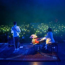 VLVV HOLDINGS - Scena koncertowa z dwoma muzykami: mężczyzna z gitarą stoi tyłem do widza, a kobieta gra na keyboardzie. W tle tłum ludzi z uniesionymi telefonami, tworzącymi efekt świetlny. Na scenie leży...