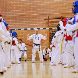 VLVV HOLDINGS - Instruktor karate demonstruje postawę przed grupą uczniów w białych kimonach i ochraniaczach na tle ceglanej ściany i siatki