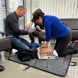 VLVV HOLDINGS - Osoby ćwiczą resuscytację krążeniowo-oddechową na manekinie z użyciem maski resuscytacyjnej i defibrylatora w pomieszczeniu z żaluzjami.