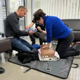 Osoby ćwiczą resuscytację krążeniowo-oddechową na manekinie z użyciem maski resuscytacyjnej i defibrylatora w pomieszczeniu z żaluzjami.