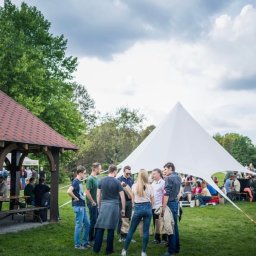 VLVV HOLDINGS - Grupa ludzi w plenerze, stojąca na trawie obok białego namiotu w kształcie gwiazdy i drewnianej altany z czerwonym dachem, w tle drzewa i pochmurne niebo.