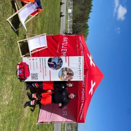 VLVV HOLDINGS - Stanowisko mobilne z czerwonym namiotem i logo 'Ratujemy+', z dwoma ratownikami w strojach oraz torbą medyczną, ustawione na trawie w słoneczny dzień, obok leżaków plażowych.