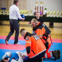 VLVV HOLDINGS - Ratownicy medyczni udzielają pierwszej pomocy zawodnikowi leżącemu na macie, widoczne trofea w tle.