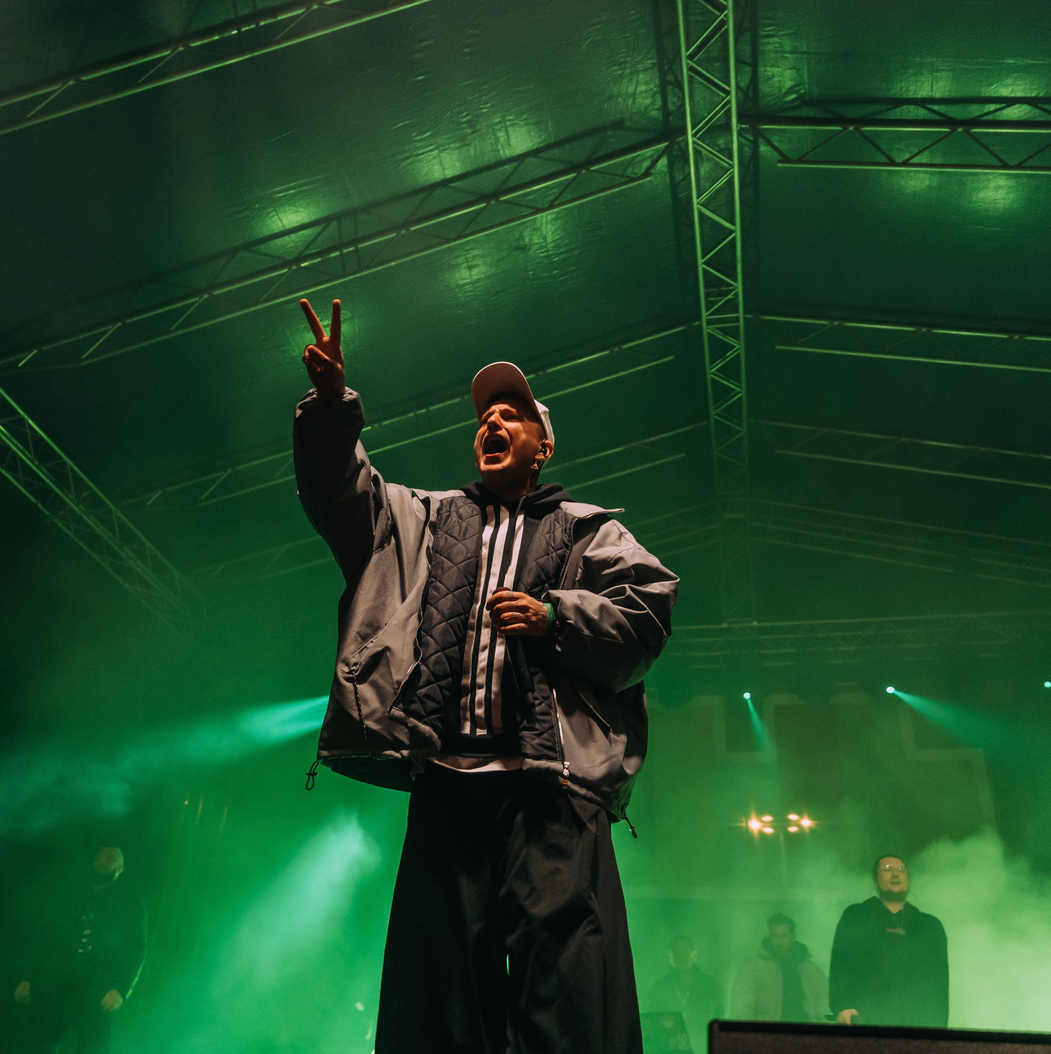 Wokalista w czapce i kurtce z trzema paskami, z podniesioną ręką i gestem V, na scenie oświetlonej zielonymi reflektorami, widoczna konstrukcja dachu scenicznego.