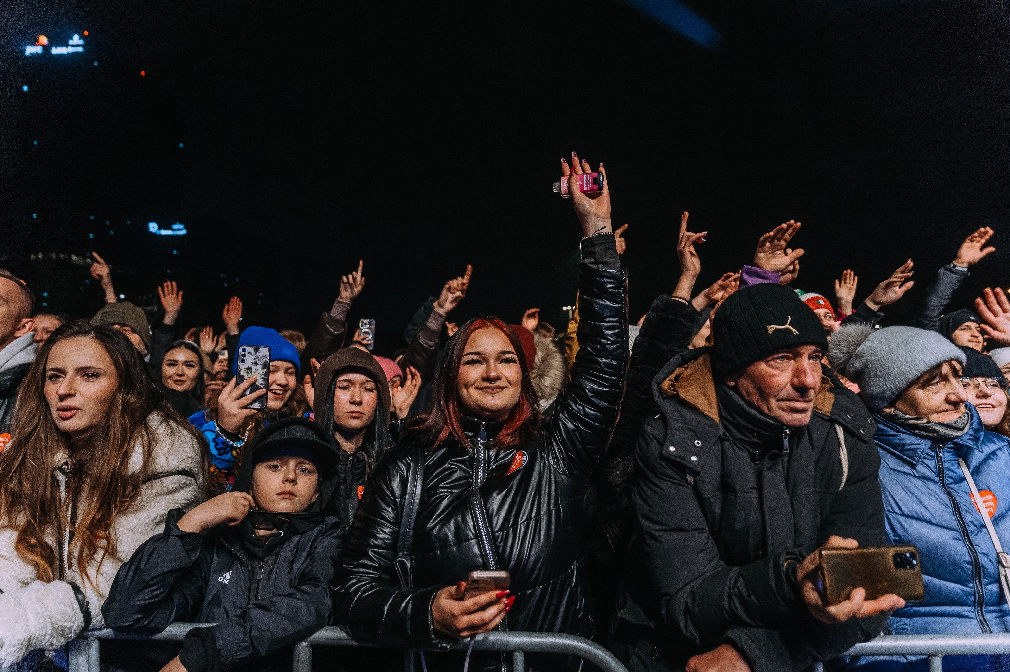 Tłum ludzi podczas koncertu plenerowego nocą, niektórzy trzymają uniesione telefony i ręce, widoczna scena w tle.