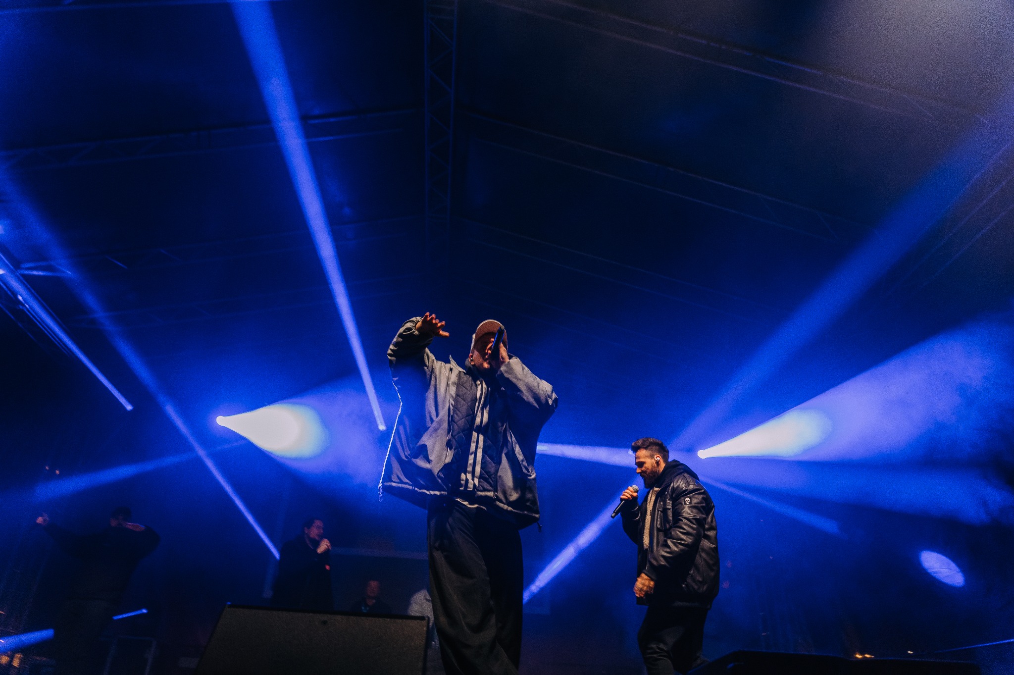 Dwóch wokalistów na scenie oświetlonej niebieskimi reflektorami, jeden z mikrofonem w ręku i podniesionymi rękami, drugi w skórzanej kurtce również z mikrofonem, w tle widoczni inni muzycy.