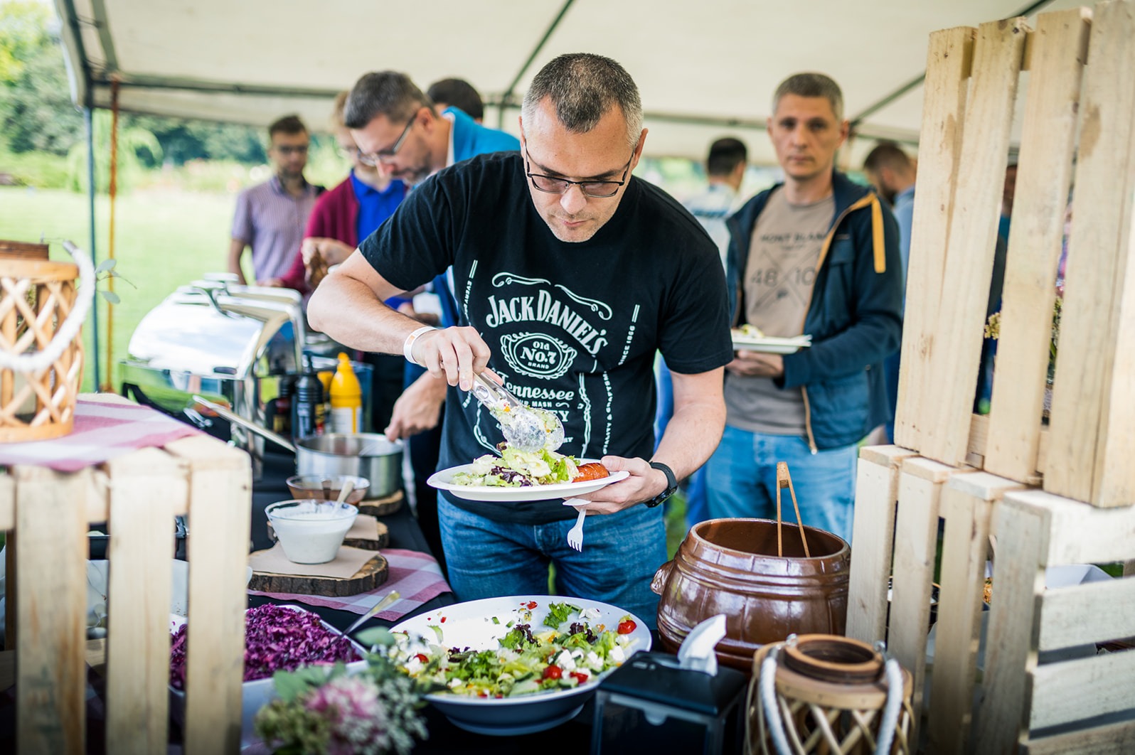 Mężczyzna w czarnej koszulce z logo Jack Daniel's nakłada sałatkę z bufetu na talerz podczas wydarzenia plenerowego, widoczny fragment stołu z różnorodnymi potrawami i dekoracjami.