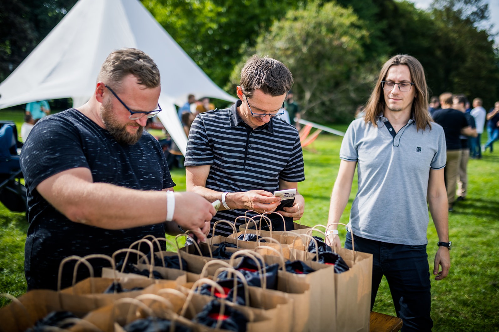 Trzech mężczyzn w okularach przygotowuje upominki w papierowych torbach na zewnątrz, widoczny biały namiot i trawnik.