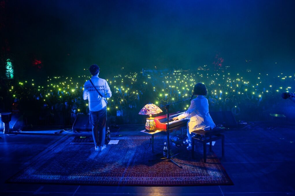 Scena koncertowa z dwoma muzykami: mężczyzna z gitarą stoi tyłem do widza, a kobieta gra na keyboardzie. W tle tłum ludzi z uniesionymi telefonami, tworzącymi efekt świetlny. Na scenie leży...
