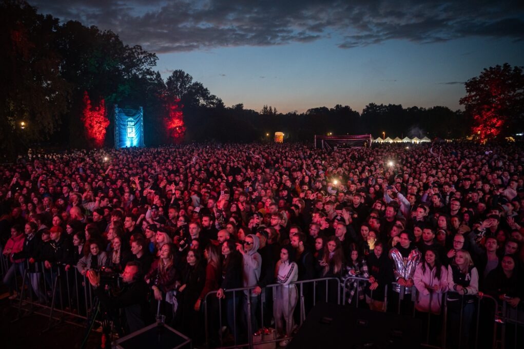 Tłum ludzi na koncercie plenerowym o zmierzchu, oświetlony czerwonymi i niebieskimi światłami sceny.