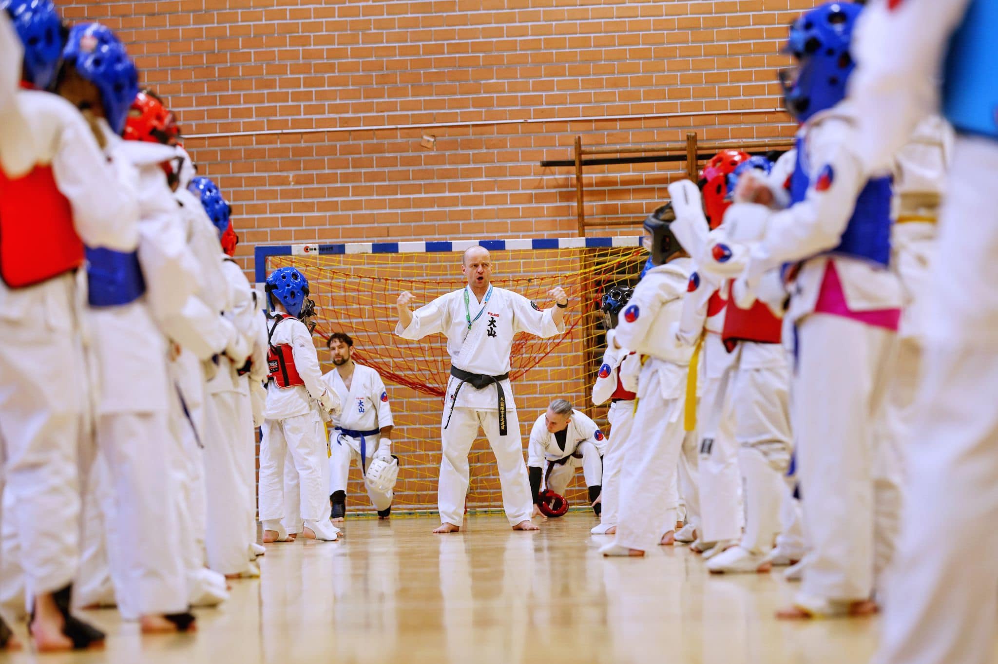 Instruktor karate demonstruje postawę przed grupą uczniów w białych kimonach i ochraniaczach na tle ceglanej ściany i siatki